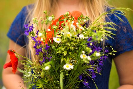 Horizontal picture of bouquet from fresh wild flowers, chamomile and poppies, in hands of young blonde girl. Unfocused face. Selective focus on flowers in hands. Side view of bouquetの写真素材