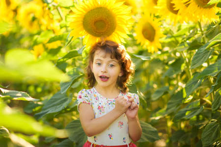 Portrait of happy surprised 4 years old curly girl in sunflowers field with big bright yellow sunflower in sunny summer day. Happy summer vacation. Close to nature childrenの写真素材