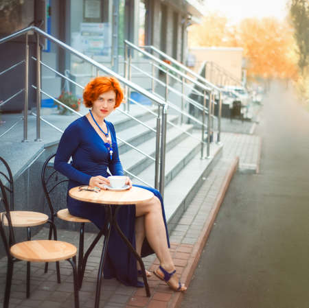 Beautiful looking red haired 30 years old woman sitting in cafe with cup of coffee. Time for yourself in summer day. Morning tea or coffee in modern street restaurant.の写真素材