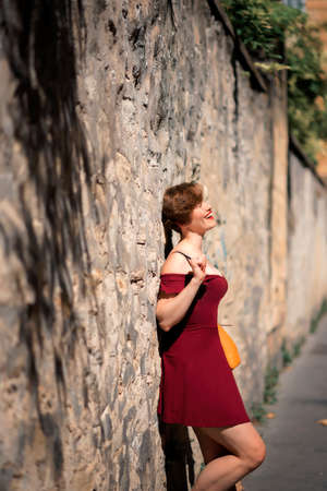 Beautiful woman in red dress standing by stone wall on summer day. Portrait of beautiful short hair woman leaning against stone wall. Tourist on vacation in Provence, France, Italy.の写真素材