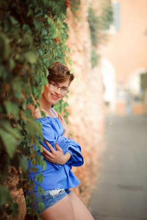 Beautiful woman in jeans shorts and glasses standing by stone wall with ivy in summer day. Portrait of short hair woman leaning against stone wall. Tourist on vacation in Provence, France, Italy.の写真素材