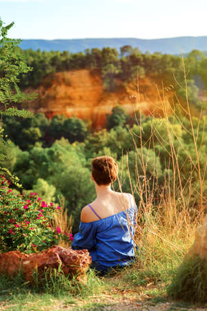 Vertical picture of short haired girl in blue shirt sitting on top of hill in sunset time and looking on ocher hills near Roussillion, Provence, France. travel tourism destination.の写真素材