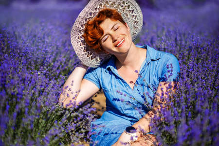 Happy red haired woman with short haircut and closed eyes in blue dress and straw hat sitting in hot summer day in lavender field. Rural summer in Provence. Travel.の写真素材