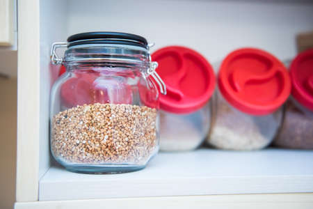 Various legumes: beans, chickpeas, buckwheat, lentils in glass jars with red covers in white cabinet. Healthy vegetarian food concept. Organizational space - management of environment.の写真素材