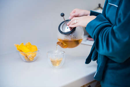 Woman's hand holding transparent kettle while pouring tea into a transparent mug. Flower tea brewed in a glass teapot with pieces of mango on table. Close up. Organic healthcare. Morning woman ritualの写真素材