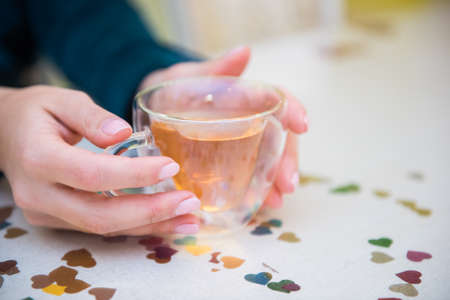 Side view of hands of a girl holding a transparent cup with green tea sitting near table. Bright highlights on a glass. Cozy picture. Close up. Organic healthcare.の写真素材