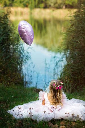 Vertical picture of back pretty little faceless blonde girl dressed pink dress sitting in the nature against the beautiful view of forest and lake. Preteen kid holding balloon. Hope and happinessの写真素材