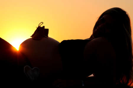 A silhouette of a pregnant woman lying on her back in front of a sunrise peaking over he stomach with a warm orange skyの写真素材