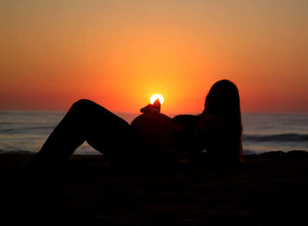 A silhouette of a pregnant woman lying on her back in front of a sunrise peaking over her stomach with a warm orange sky and ocean backgroundの写真素材