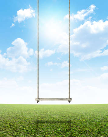 A regular home made swing made of rope and a wooden plank on a grassy field ground and blue cloudy sky backgroundの写真素材