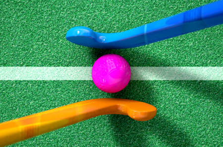 A 3D rendering of two hockey sticks stadning opposite each other over a ball on green artificial grass in the daytimeの写真素材