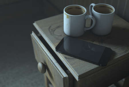 A real life concept scene showing a bed side table with dirty coffee mugs and a generic smartphone at night - 3D Renderの写真素材