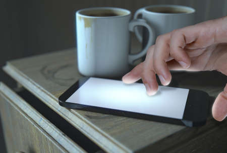 A bed side table with coffee mugs and a generic illuminated smartphone and a hand touching it - 3D Renderの写真素材