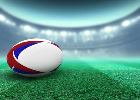 A reguar white rugby ball with red and blue design elements resting on a stadium grass pitch at night under illuminated floodlights - 3D renderの写真素材