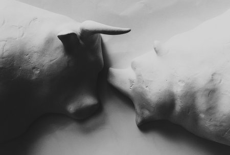 Two white clay statuettes depicting a stylized bull and bear in dramatic light representing...