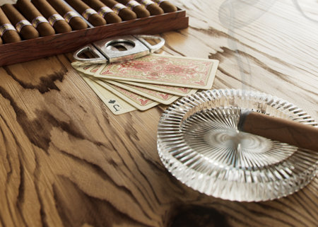 A scene with a set of cigars and antique playing cards and a lit cigar in an ashtray on a wooden surface - 3D renderの写真素材