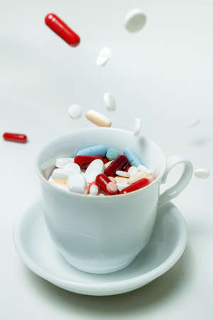 A cup of coffee with medicines of different shapes and colors inside on a white background. Capsules and pills fall into a mug on a white desk.の写真素材