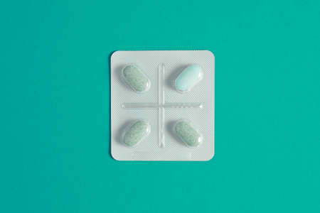 A square blister pack with four pills and two lines in the shape of a cross on a blue background. Three green and one cyan pills inside transparent plastic on a table.の写真素材