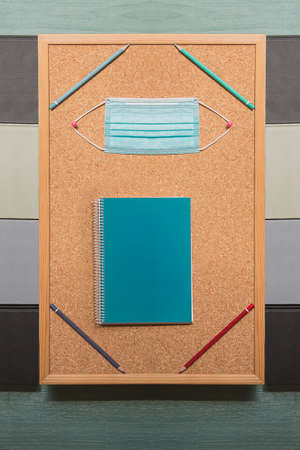 A corkboard with colored pencils in its four corners supports a surgical mask held by two pink pins...