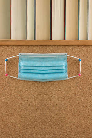 A surgical mask placed on a cork board with a row of books on top and space below to put a text.の写真素材