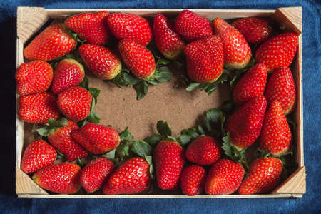 Overhead view of a wooden tray with a copy space in the middle surrounded by fresh strawberries on a blue fabric texture background. Healthy and vegetarian food.の写真素材