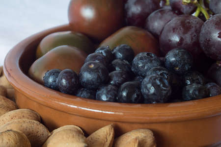 Close Up of some blueberries with a bunch of black grapes, tomatoes, and almonds in a clay bowl on a kitchen table in a home. Nuts, vegetables, and fruit. Healthy food.の写真素材