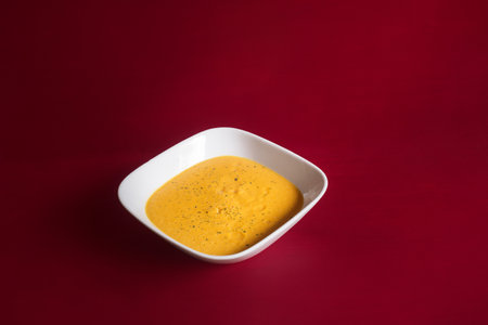 A plating of a delicious vegetable cream with crushed carrot and pumpkin is presented in a white ceramic bowl on a red-painted wooden table with copy space. Healthy food.の写真素材