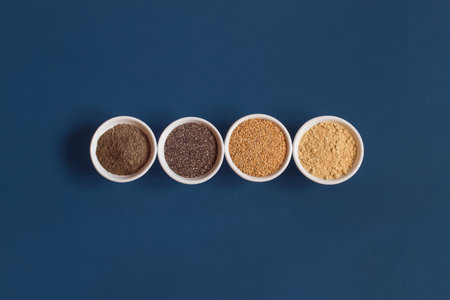 Four white ceramic bowls in a row with both whole and powdered chia and flax seeds on a blue studio...
