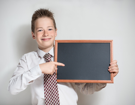 The nice smiling boy teenager in a white shirt and a tie on a grey background specifies a finger in an empty black board for a chalkの写真素材