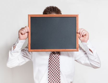 The boy in a white shirt and a tie holds empty black chalkboard,with place for text,closing his faceの写真素材