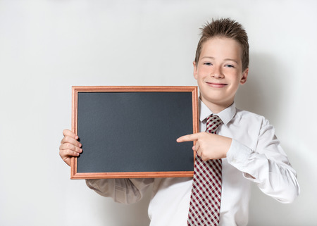 The nice smiling boy teenager in a white shirt and a tie on a gray background specifies a finger in an empty black board for a chalkの写真素材