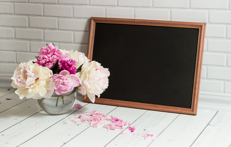 Beautiful bouquet of pink and white peonies in the glass vase with petals and black chalkboard with empty place for your text on the light grey brick and wood background.の写真素材