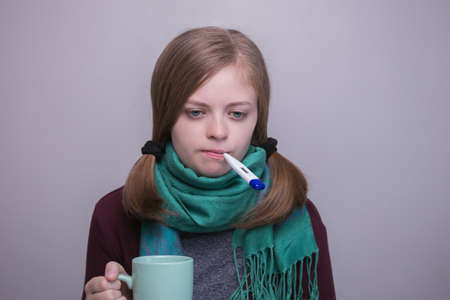 Young caucasian woman is sick and keeps a thermometer in her mouth and cup in hands