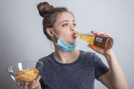 Smiling young caucasian woman girl in mask drinking bottle of beer and holding potato chips crispsの写真素材