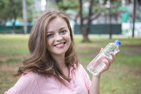 Smiling young caucasian woman girl holding drinking water from a plastic bottle in green parkの写真素材