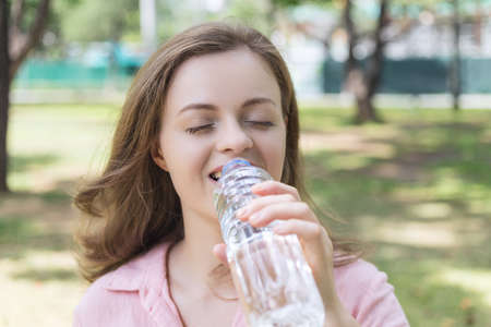 Young caucasian woman girl drinking water from a plastic bottle in green park, close up