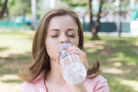 Young caucasian woman girl drinking water from a plastic bottle in green park, close up