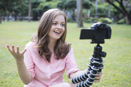 Smiling young caucasian girl woman making a video blog (vlog) with camera outside in green parkの写真素材