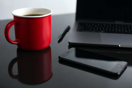 Stylish red coffee cup, phone and laptop on desk table. Black background, business concept, close upの写真素材