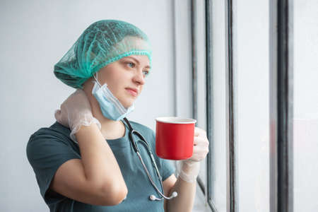 Young caucasian female doctor nurse in surgical mask looks tired. Coffee break during coronavirus covid-19 outbreakの写真素材