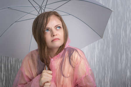 Young Caucasian woman girl in a pink raincoat holding umbrella under heavy rain with funny annoyed...
