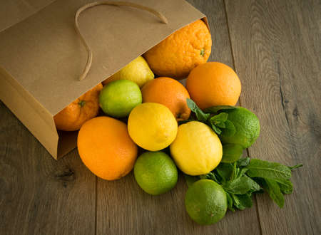 Paper bag with fresh citrus on brown wooden background. Many fruits falling out from package. Pile of juicy oranges, lemon, lime, mint. Grocery store. Buying natural organic products. Vegetarian foodの写真素材