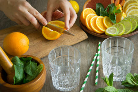 Female hand cutting orange pieces on wooden board, making fresh summer citrus cocktail, lemonade. Sliced orange, lemon, grapefruit, lime, mint on plate. Mortar, pestle, glasses, straw on foregroundの写真素材