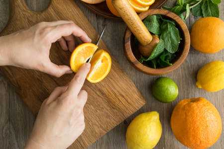 Top table view of female hands cutting orange pieces on wooden board, preparing fresh summer citrus sweet food. Solid orange, lemon, grapefruit, lime, and mortar with pestle, mint staying aroundの写真素材