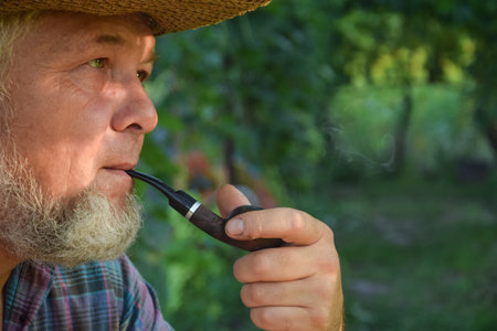 Senior adult man with white beard smoking tobacco pipe outdoors. Countryman wearing straw hat. Male villager looking thoughtfully at garden. Contemplation, relaxingの写真素材