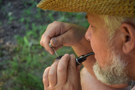 Senior adult man with white beard smoking tobacco pipe outdoors. Countryman wearing straw hat. Male...