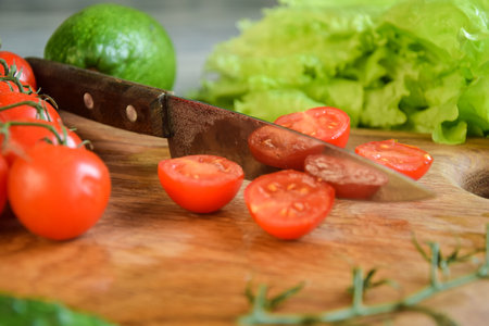 Closeup kitchen knife, sliced tomatoes on wooden cutting board on background of green vegetables indoors. Food cooking background. Ingredients for preparation salad dishes. Healthy food conceptの写真素材