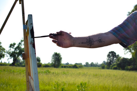 Artist's hand with tattoo paints canvas by paintbrush on wooden easel. Golden Sun on Blue Skyline Horizon. Modish middle-aged painter creating artwork at field sunrise. Scenic landscape nature viewの写真素材