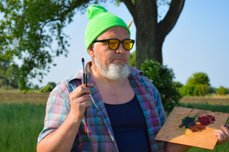 Enthusiastic painter working outdoors having creative inspiration holding paintbrush and palette with oil paints or acrylic. Mature man artist hipster creating artwork oil painting at meadow dawnの写真素材