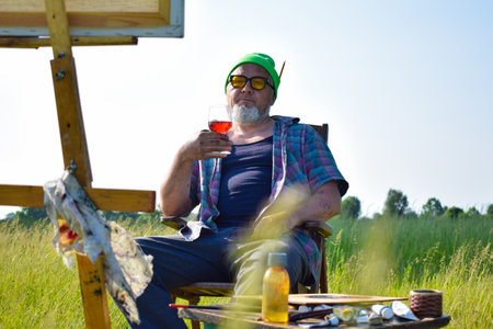 Offbeat mature artist relaxing after completion artwork at field, drinks wine looking at picture sitting on chair outdoors in morning sunshine. Canvas on wooden easel painting tools materials suppliesの写真素材
